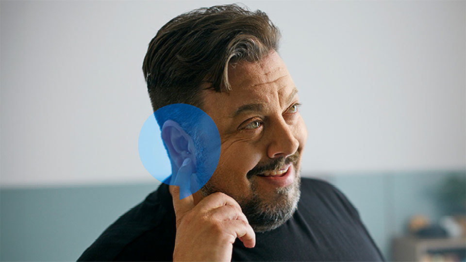 A person's ear, marked by a blue highlight, is centered while a hand gestures nearby in a modern indoor setting, suggesting hearing care or hearing aid fitting in an Audika clinic. No visible logos or text.