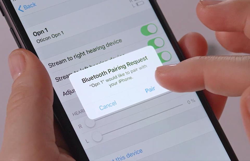 A smartphone screen displays a Bluetooth pairing request for an ’Oticon Opn 1’ hearing aid. A hand selects ’Pair’ in a hearing clinic or setup environment.