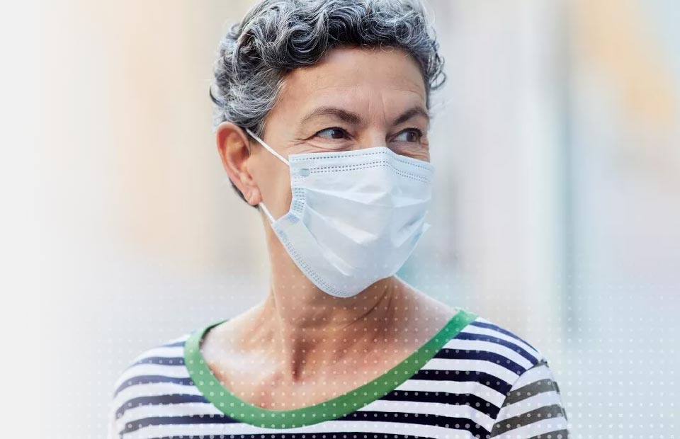 An individual with short, curly gray hair wears a striped shirt with green trim and a medical mask, set against a softly blurred indoor or outdoor environment with warm lighting.