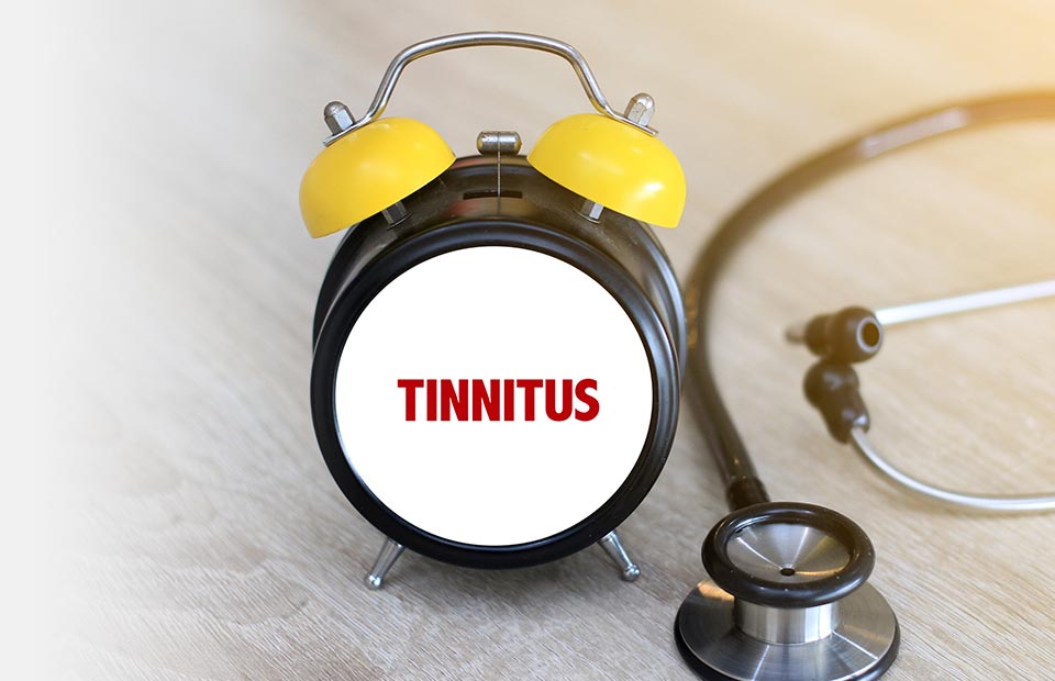 A black analog alarm clock with yellow bells displays the word ’TINNITUS’ in bold red letters on its face. Beside it, a stethoscope rests atop a wooden surface, evoking a hearing clinic or healthcare context.