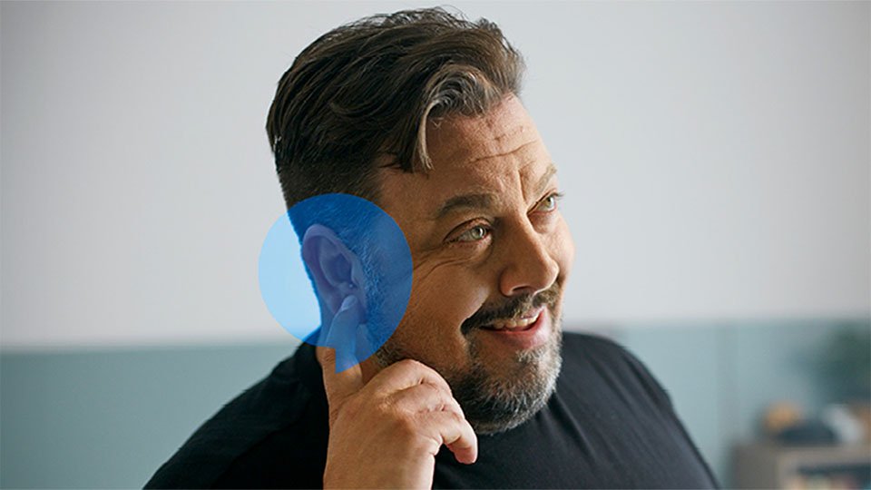 A person’s ear, marked by a blue highlight, is centered while a hand gestures nearby in a modern indoor setting, suggesting hearing care or hearing aid fitting in an Audika clinic. No visible logos or text.