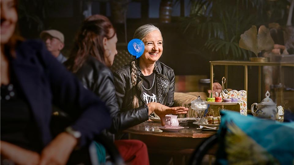 Two individuals converse at a cozy café table adorned with teacups, macarons, and a teapot. The blurred central figure wears a prominent blue earmuff-like object, suggesting hearing-related imagery.
