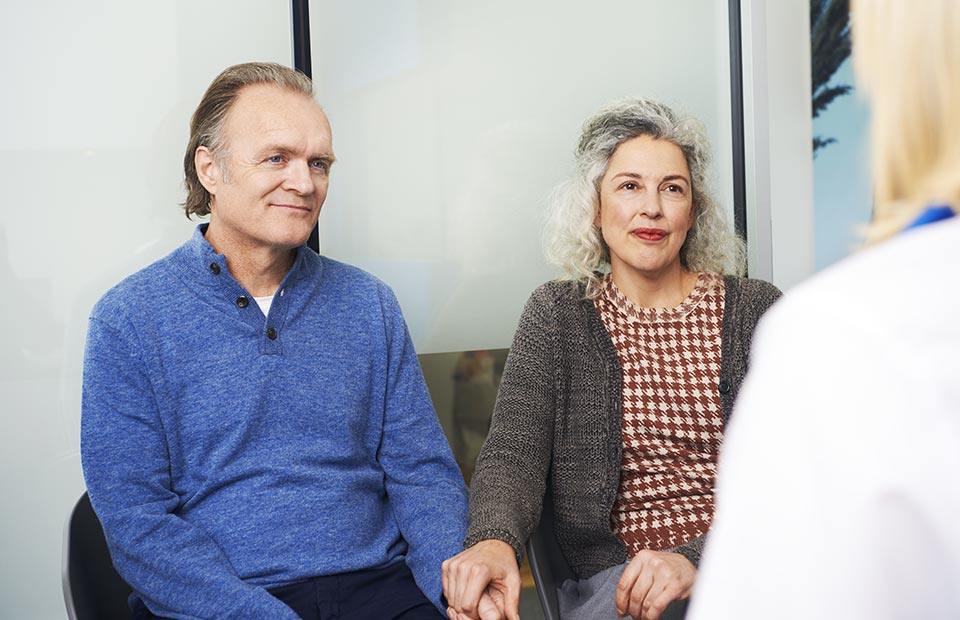 A middle-aged couple sits holding hands during a consultation with a hearing care professional at a clinic. The setting includes bright, modern interiors with frosted glass panels. No visible text.