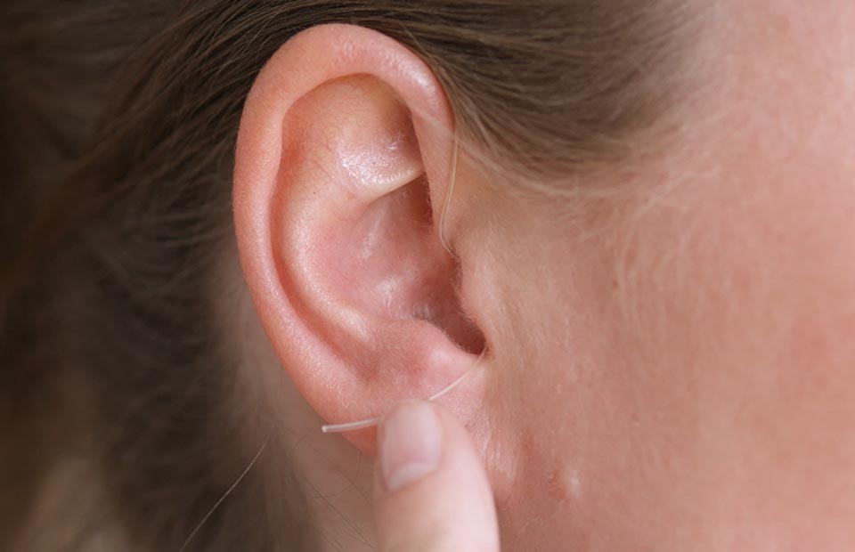 A close-up of an ear with fingers adjusting a nearly invisible, thin wire of a discreet hearing aid. The device highlights modern hearing solutions, ideal for a hearing clinic context.