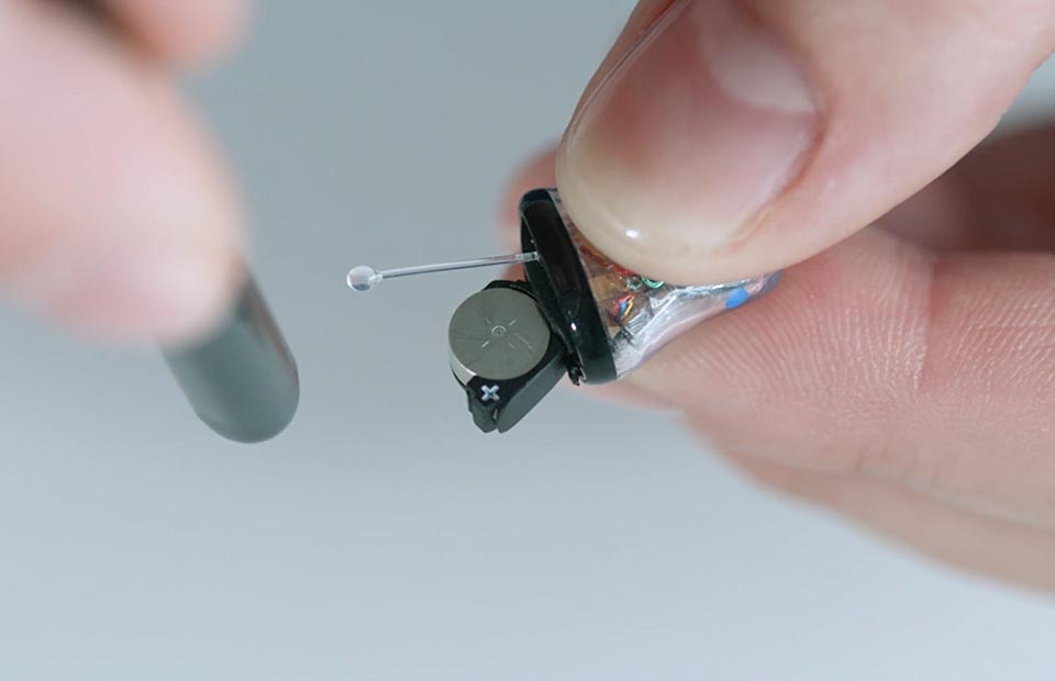 A close-up of hands handling a compact, in-the-ear hearing aid with exposed components, featuring a small battery. The setting appears clinical or instructional, emphasizing hearing care precision. No visible branding or text.