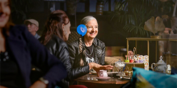 Two women in a cozy café converse over tea and desserts, surrounded by eclectic decor and plants. One wears a hearing aid, likely from Audika’s range of hearing solutions.