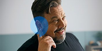 A person points to their ear with a blue overlay highlighting the ear anatomy. The setting appears to be indoors, possibly a hearing clinic, with a blurred background. No visible text.