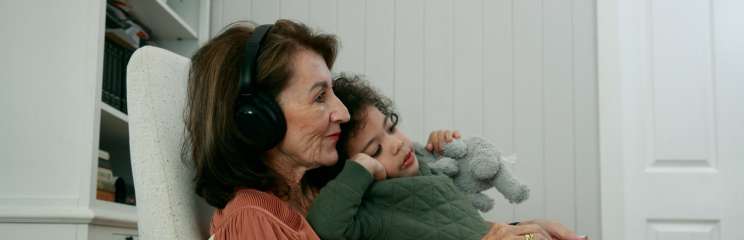 A woman wearing headphones holds a child clutching a stuffed elephant in a cozy, home-like environment with white-paneled walls and shelves with books in the background. No visible text.