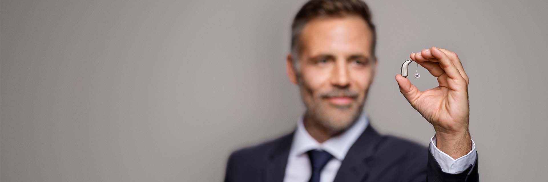 A person in formal attire holds a discreet behind-the-ear hearing aid with a thin wire, set against a plain gray background, emphasizing hearing care technology and professional expertise.
