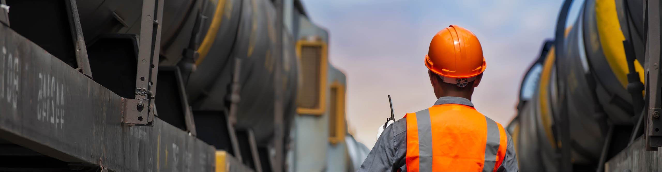 A worker in an orange safety vest and hard hat holds a walkie-talkie, standing between large freight train cars at an industrial site under a partly cloudy sky.
