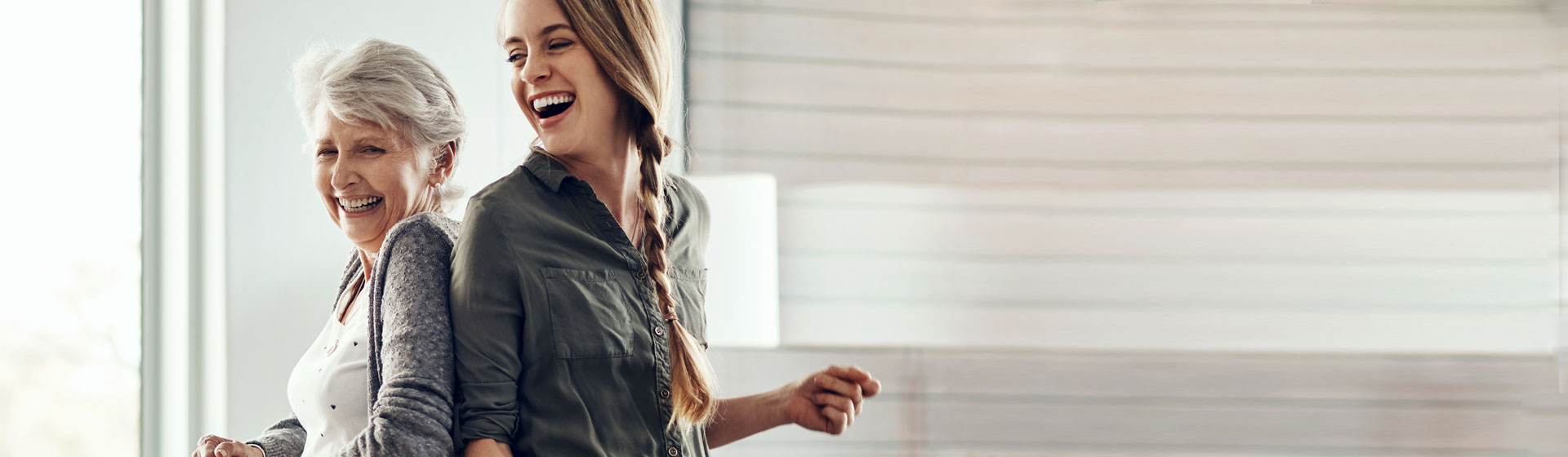 A caregiver supporting an elderly woman in a bright living space.
