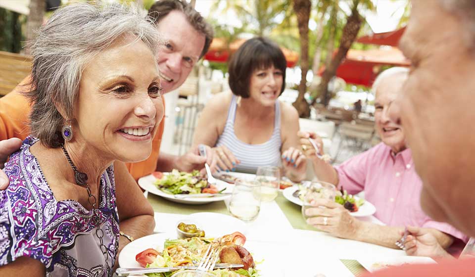 A group of people enjoys a meal together at an outdoor restaurant with tropical surroundings, including palm trees and red awnings. Plates contain salads and various colorful dishes. No visible text or identifiable hearing aids.