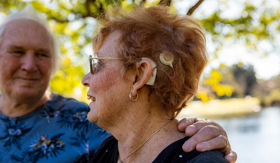An older woman with short red hair is wearing a cochlear implant on her left ear, while standing outdoors near a serene park with sunlit trees and water in the background.