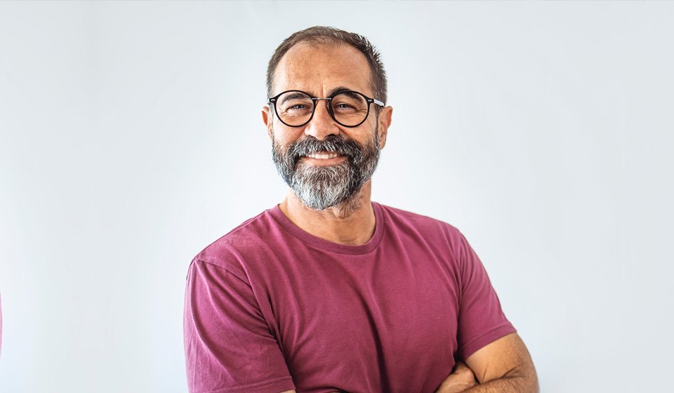A middle-aged man in a maroon t-shirt stands with arms crossed against a plain white background. The face is blurred, and no hearing aids, text, or specific brand elements are visible.