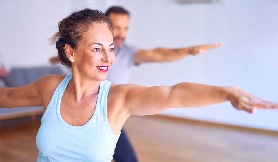 A woman in a light blue tank top holds a yoga pose, extending her arms outward in focus, with another person behind her. The setting is a bright indoor studio.