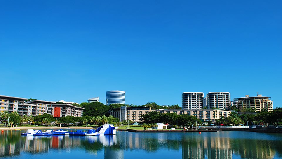 Waterfront view of modern buildings, lush greenery, and an inflatable water park under a clear blue sky.