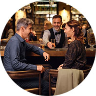 A man and woman sit conversing in a lively restaurant or bar setting, with a bartender in the background serving drinks. Surroundings feature warm lighting and a social atmosphere. No text visible.