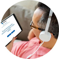 A person holds a tablet displaying an Audika webpage with the text: ’Do you have hearing issues? Find your hearing in 3 minutes with our online hearing test.’ The background shows a home setting with a beige couch.