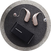 Two sleek beige hearing aids with transparent earpieces rest beside a compact black charging case on a circular gray surface, emphasizing advanced hearing solutions in a modern context. No text visible.