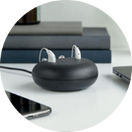 Modern rechargeable hearing aids in a sleek black charging case placed on a desk near a smartphone and laptop.