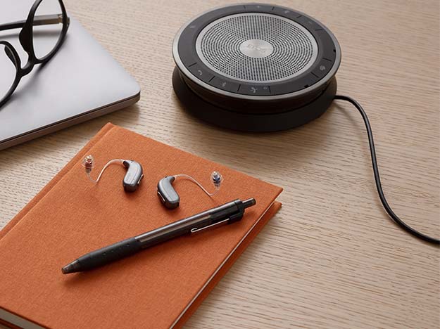 A black, circular speakerphone with a wired connection is placed on a light wooden desk alongside glasses, a closed laptop, and orange notebooks, suggesting a professional or office setting.