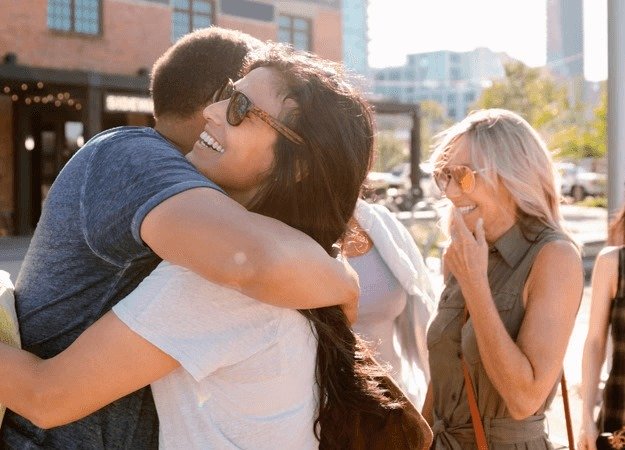 Two individuals embrace outdoors in a sunlit urban area, surrounded by buildings and greenery. Another person stands nearby, observing the scene while holding a hand near their face.