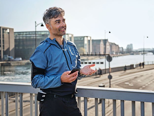 A man in a blue jacket is standing on a bridge, holding a smartphone and gesturing with his other hand near a waterfront urban setting under daylight. No visible hearing aids or text.