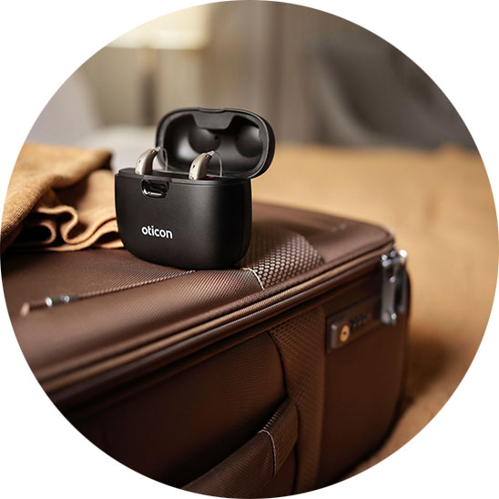 A pair of Oticon hearing aids rests in an open black charging case placed on a brown suitcase, in a softly lit bedroom with folded linens in the background.