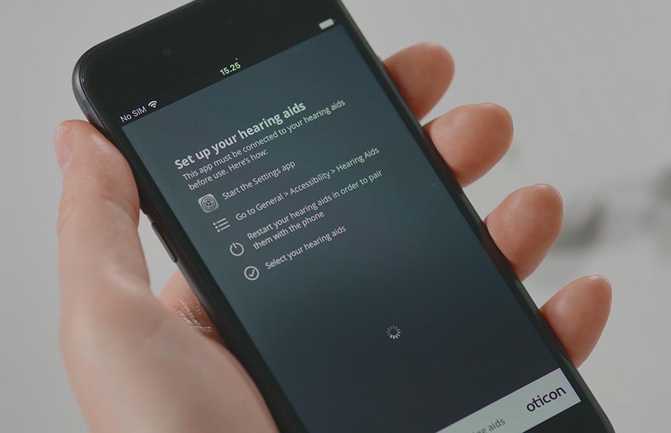 A hand holds a smartphone displaying setup instructions for hearing aids. The screen lists steps: 'Start the Settings app,' 'Go to General > Accessibility > Hearing Aids,' 'Restart your hearing aids,' and 'Select your hearing aids.' The Oticon logo is visible in the bottom-right corner, linking the image to Oticon hearing aids. The neutral, bright background suggests a hearing clinic or home setting.