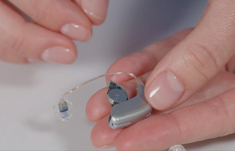 A small, sleek silver hearing aid is held delicately by hands with neatly manicured nails. The device’s battery compartment is open, showcasing its functionality within a clinical environment.