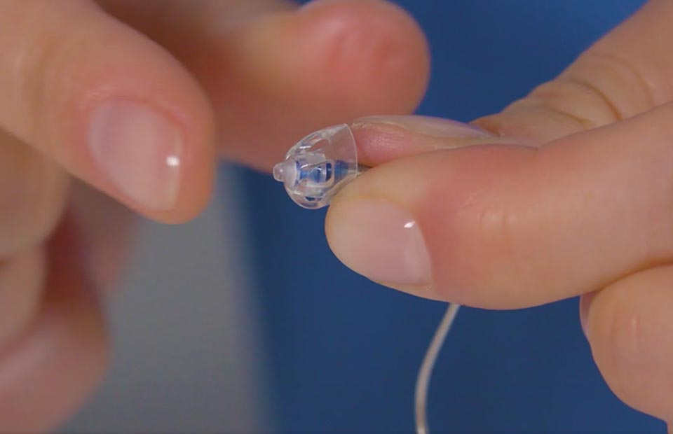 A close-up of hands holding a small, modern hearing aid with a clear dome tip, set against a blue background, emphasizing audiology care and hearing clinic expertise. No visible branding.