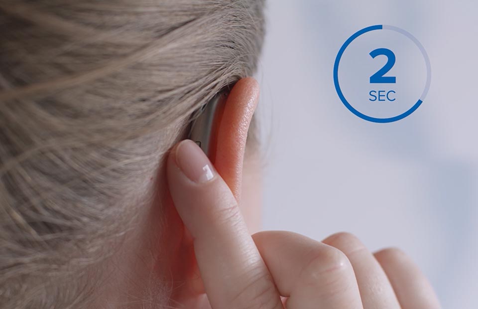 A close-up of a person adjusting a behind-the-ear hearing aid with their fingertip. The text '2 SEC' inside a blue circular graphic indicates a timed action. Background is softly blurred.