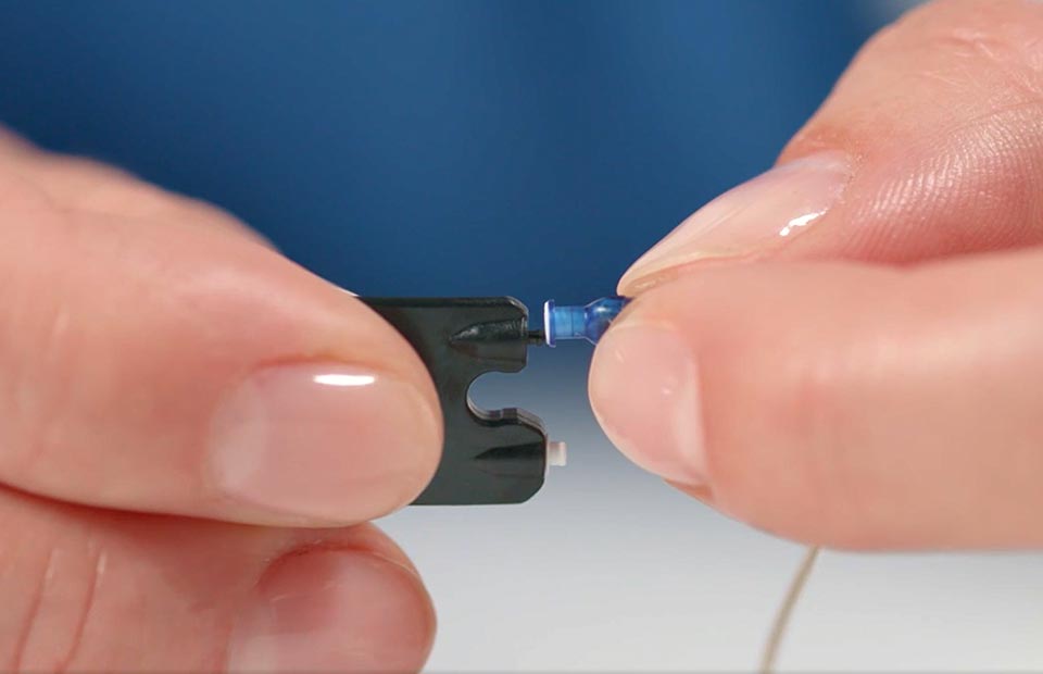 Hands hold a small hearing aid wax guard tool with a blue wax filter being inserted or replaced. The tool and filter are set against a blurred blue background, illustrating hearing aid maintenance.