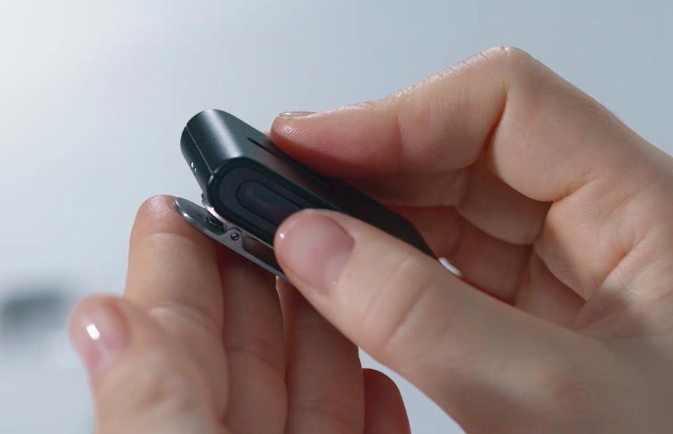 Hands hold a sleek black hearing aid accessory, likely from Oticon or Bernafon, adjusting its clip mechanism. The setting is minimalistic with a blurred, light background, emphasizing modern hearing technology.