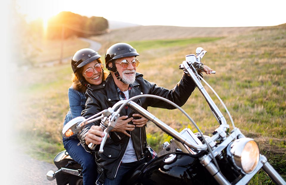 A couple rides a black motorcycle on a winding rural road at sunset, surrounded by open fields. Both wear helmets and leather jackets, enjoying a scenic, carefree experience. No visible text.