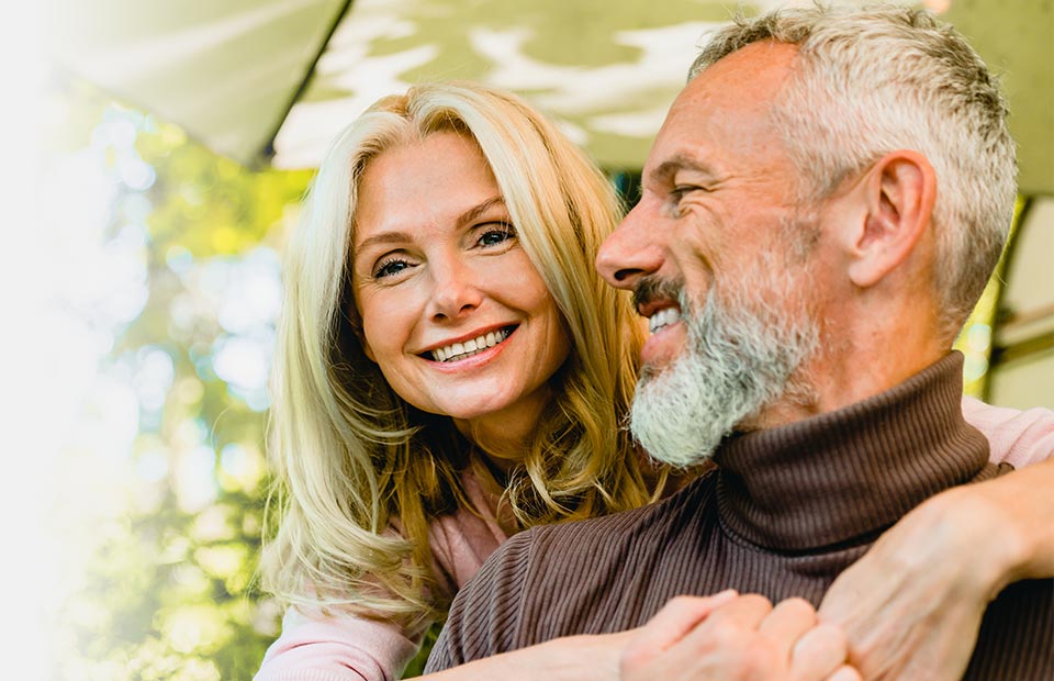 A close-up of an older couple outdoors under a patio umbrella surrounded by greenery, where the man appears to wear a subtle, skin-tone hearing aid behind his ear.