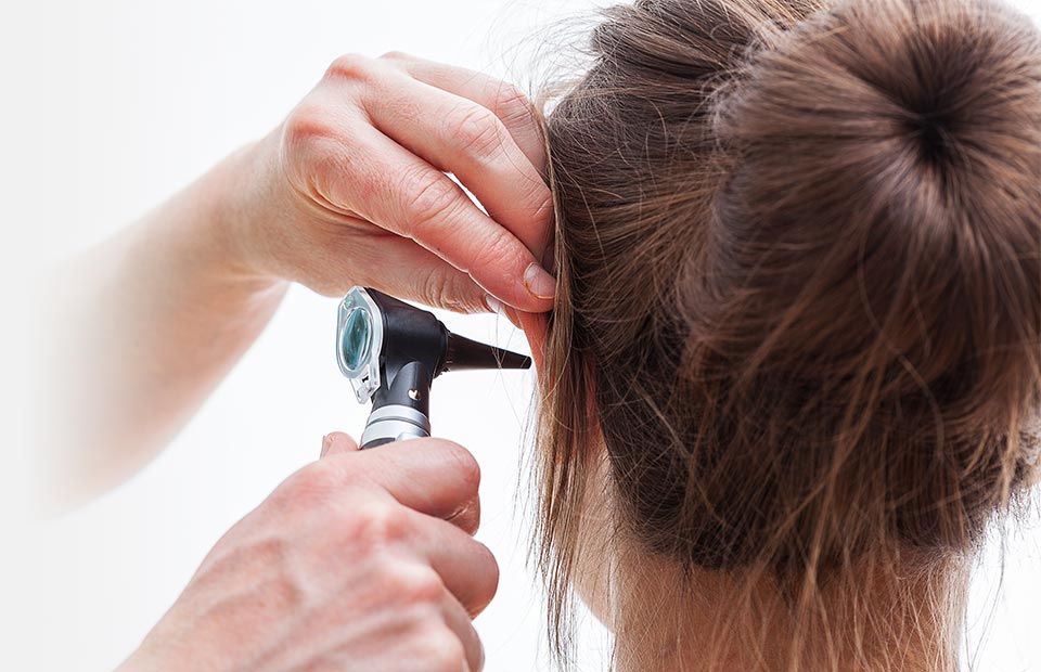 An audiologist examines a patient’s ear using an otoscope in a clinical setting, likely part of a hearing test or hearing care consultation at a hearing clinic. No text visible.