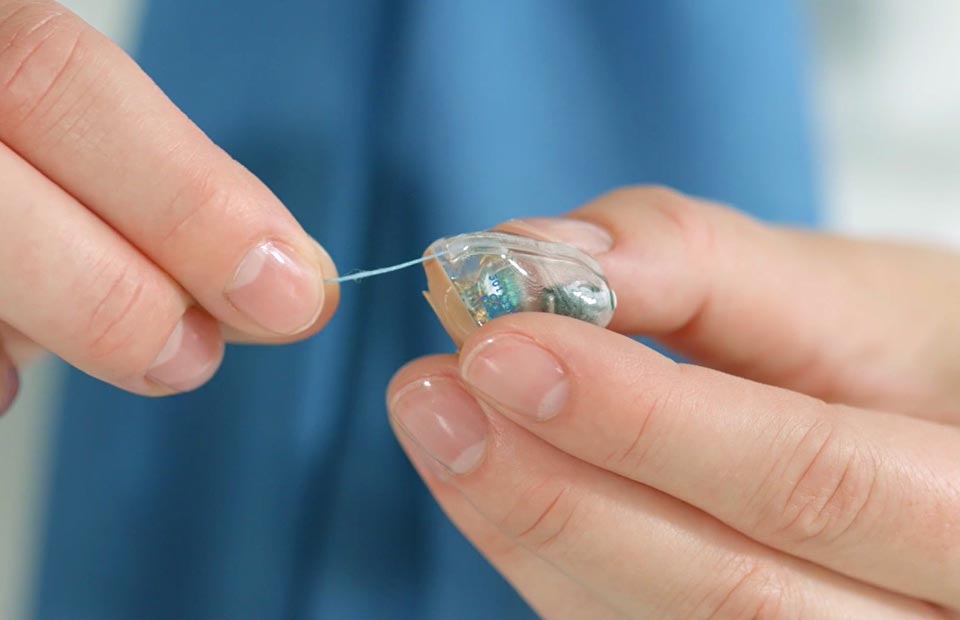 Hands hold a small, clear in-the-ear hearing aid with a removal string visible, set against a blurred blue background, likely in a hearing clinic or audiology care environment.