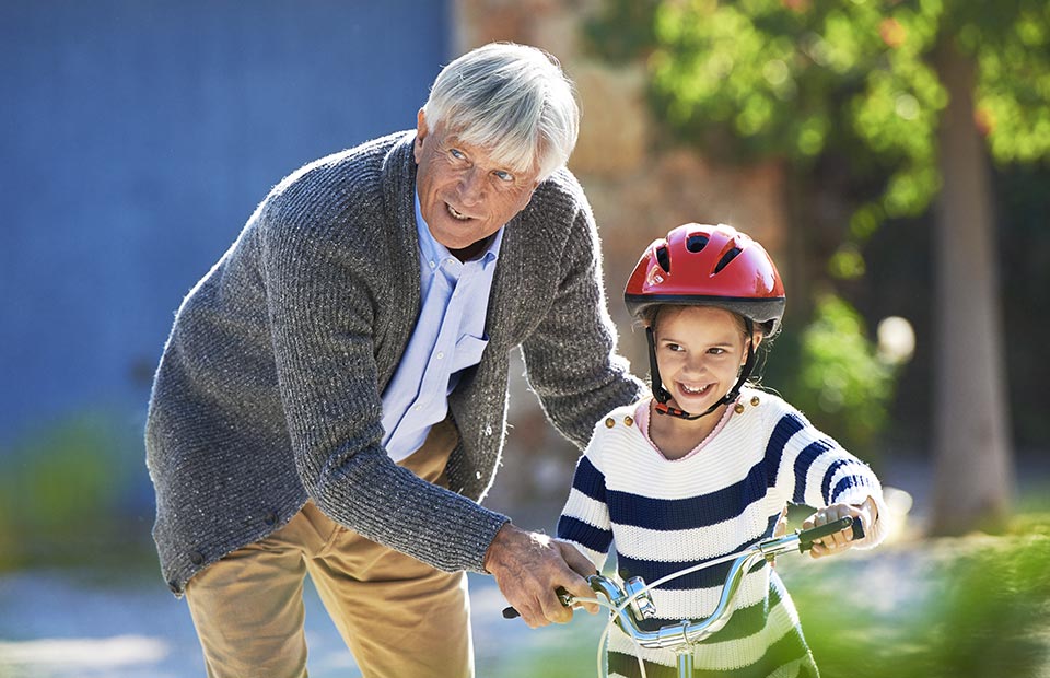 An elderly man in a grey cardigan helps a young child wearing a red helmet balance on a bicycle outdoors, surrounded by greenery and a building with a blue wall.