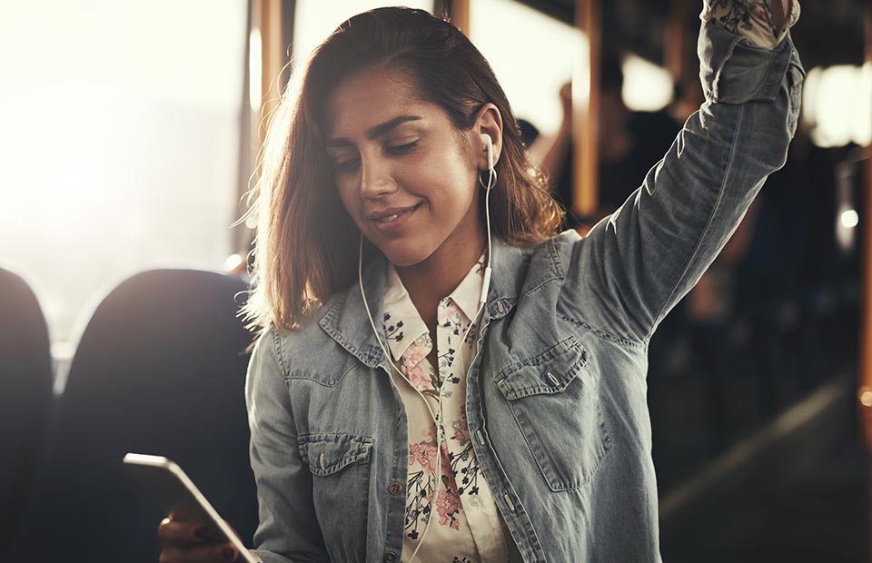 A person wearing earphones listens to audio while holding a smartphone in one hand and grasping a handrail in the other, seated in a sunlit public bus environment.