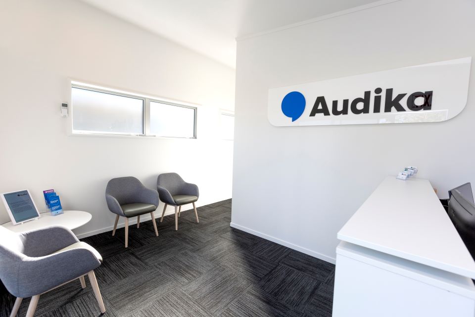 Modern hearing clinic reception area with Audika branding, gray seating, white furniture, and large windows for natural light.