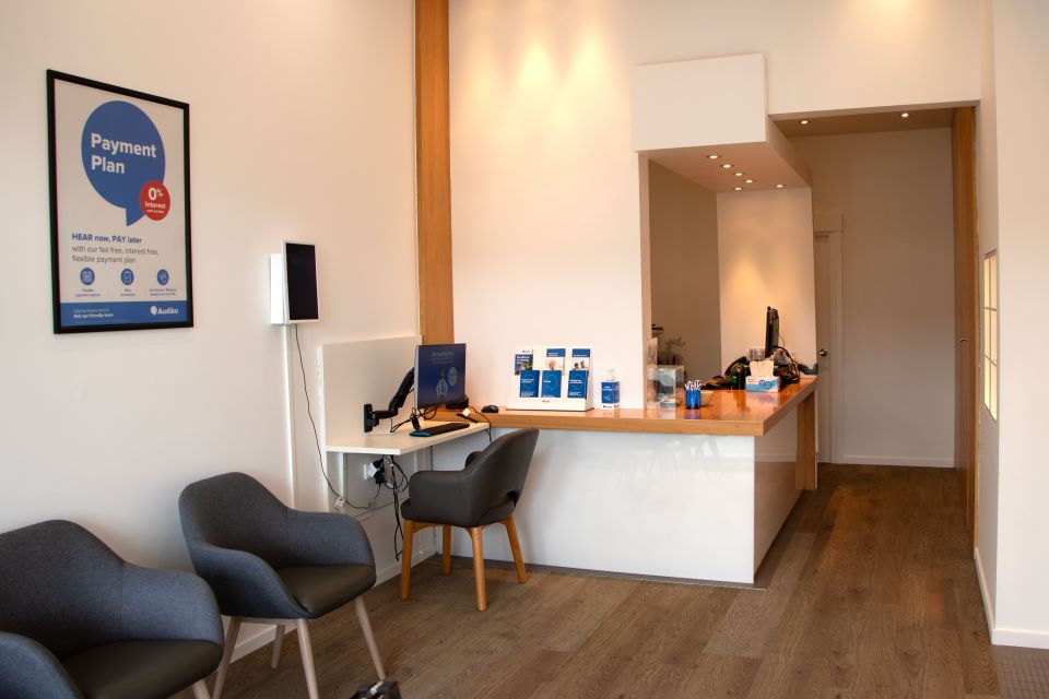 Hearing care clinic reception area with modern seating, a wooden desk featuring hearing care literature, and a sign promoting a payment plan option.