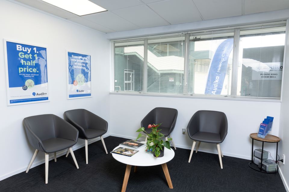 Waiting area at Audika Hamilton featuring modern gray chairs, a white coffee table with magazines and a plant, promotional posters on the wall, and natural light from large windows.