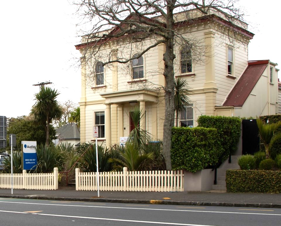 Audika Hearing Aid Clinic in Remuera, featuring a historic cream-colored building with lush greenery and a white picket fence by the roadside.