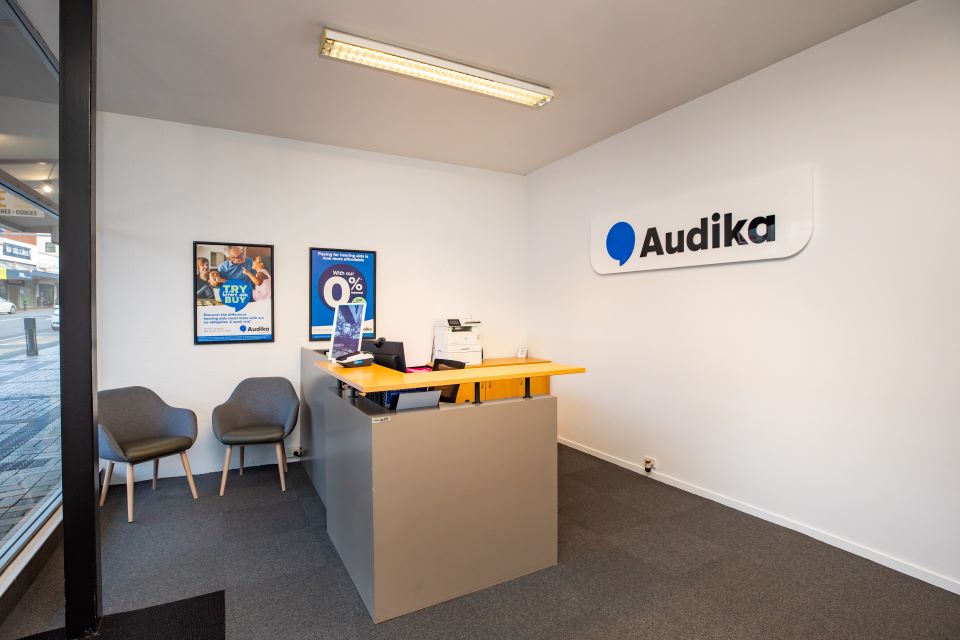 Reception area of Audika hearing aid clinic in Timaru featuring a desk, chairs, wall posters, and the Audika logo.