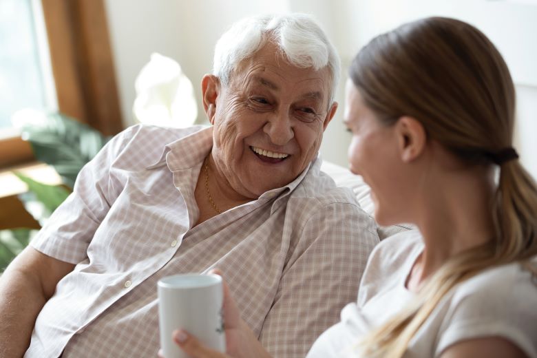 An elderly man and a younger woman converse while seated in a well-lit room with houseplants near a window. The woman holds a white mug, suggesting a casual, intimate discussion.