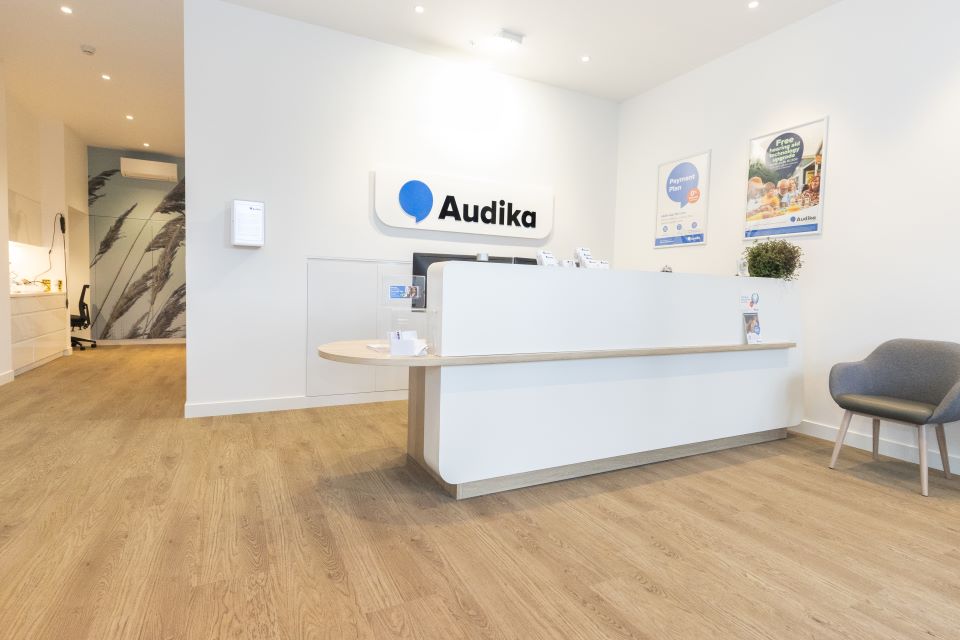 Front desk and reception area of Audika Barrington hearing care clinic with modern white decor, wooden flooring, and informational posters on display.