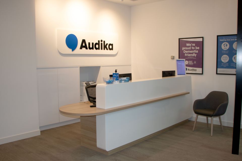 Audika Mission Bay reception area with modern white decor, a welcoming desk, branded signage, and informational posters emphasizing dementia-friendly services.
