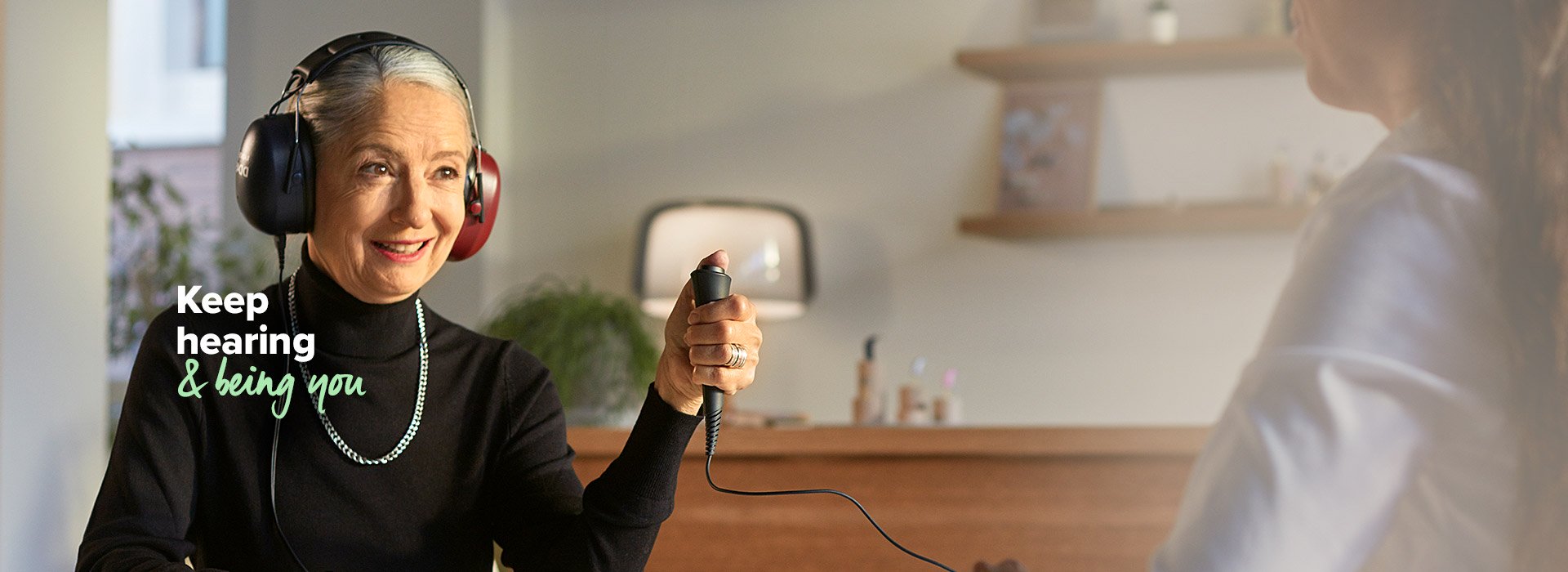 Senior woman wearing headphones during a hearing test while holding a response button, with text overlay ’Keep hearing & being you’.