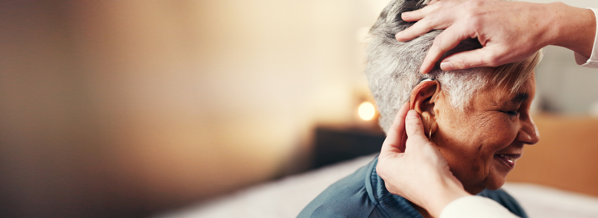 Lady_with_short_grey_hair_turned_side_on_being_assisted_by_an_audiologist_with_help_fitting_her_hearing_aid